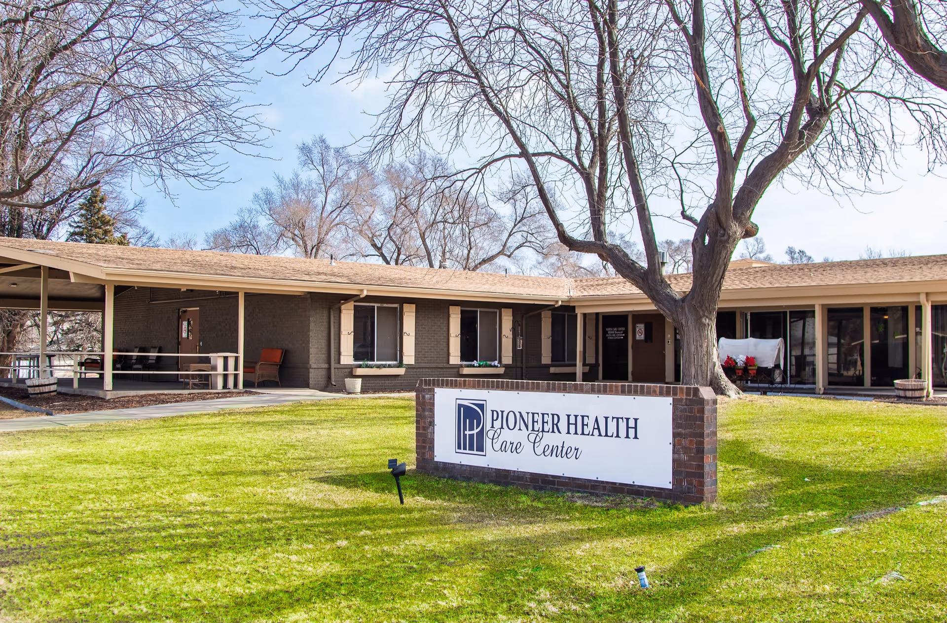 Exterior view of Pioneer Health Care Center, a single-story building with a covered walkway, large windows, and a sign on the lawn displaying the facility's name. Leafless trees are visible in the background under a partly cloudy sky.