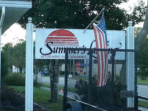 Outdoor sign for Summer's Landing Retirement Community with a red sun graphic, an American flag attached to a black metal fence in front of the sign, and trees and a street in the background.