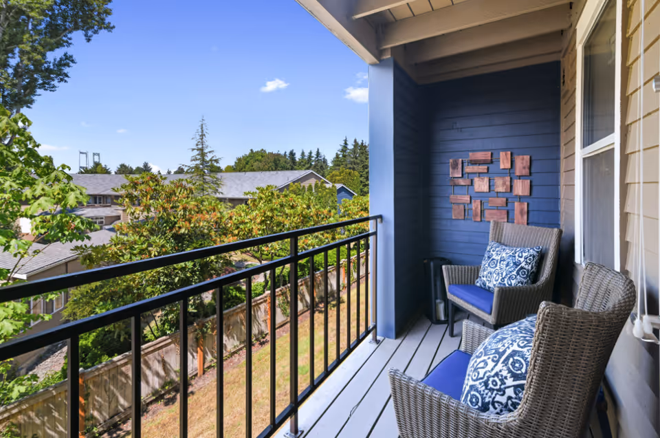 A small balcony with two wicker chairs featuring blue cushions and patterned pillows. The balcony has a black metal railing and overlooks a residential area with trees and houses under a clear blue sky. There is a decorative wooden wall art piece on the blue wall of the balcony.