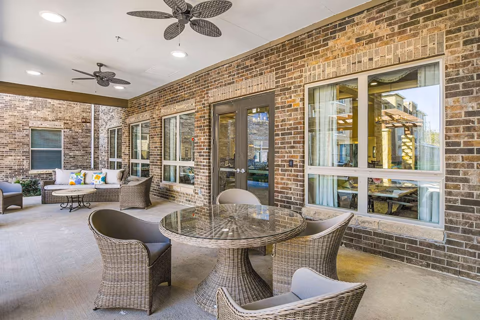 Covered brick patio with a round wicker dining table and chairs and a cushioned seating area by large windows and double doors.