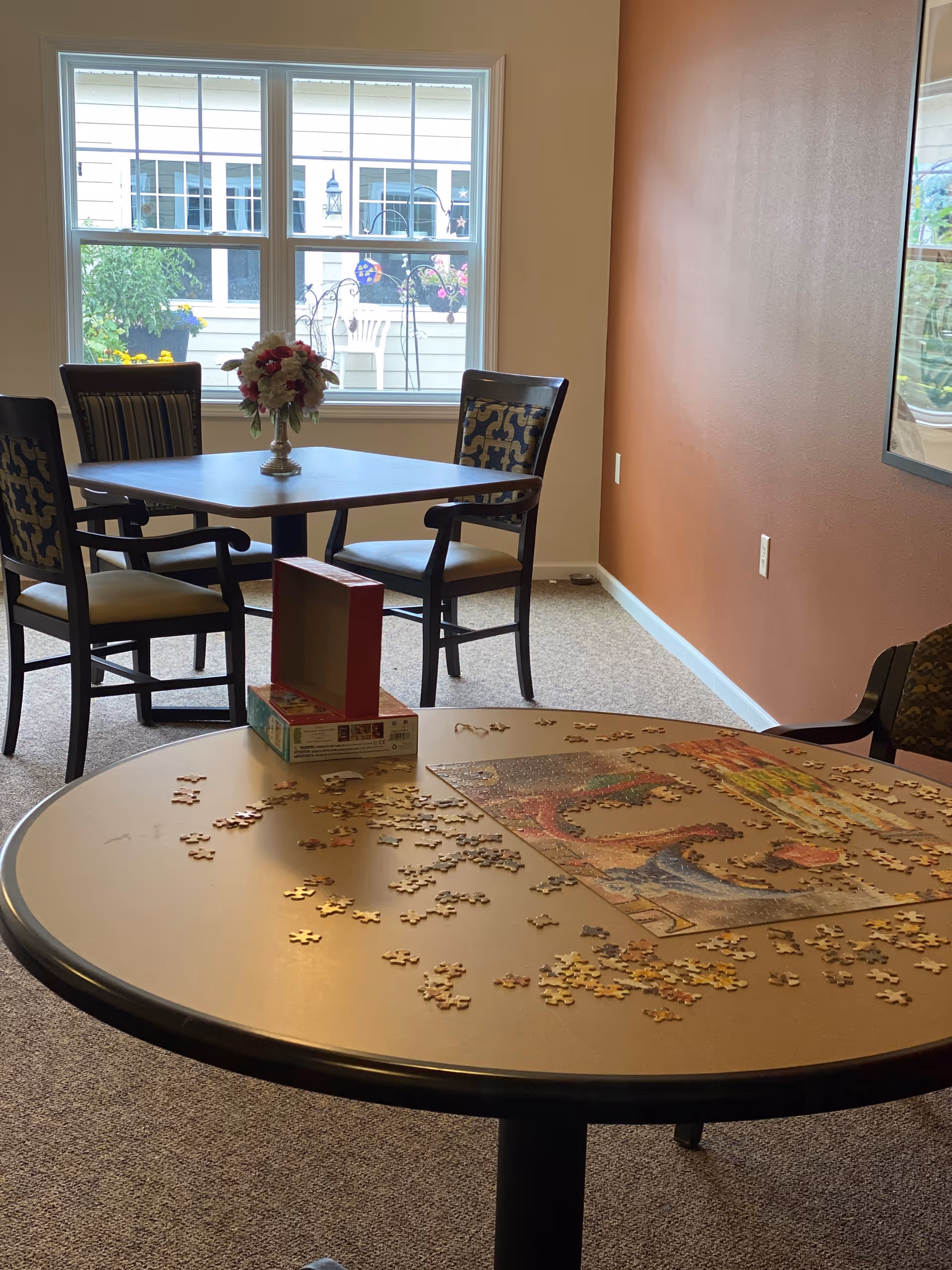 A cozy room with two tables and several chairs. The foreground table has a partially completed jigsaw puzzle with scattered puzzle pieces and an open puzzle box. The background table has a vase with flowers and is positioned near a window showing an outdoor view with plants and a white chair.