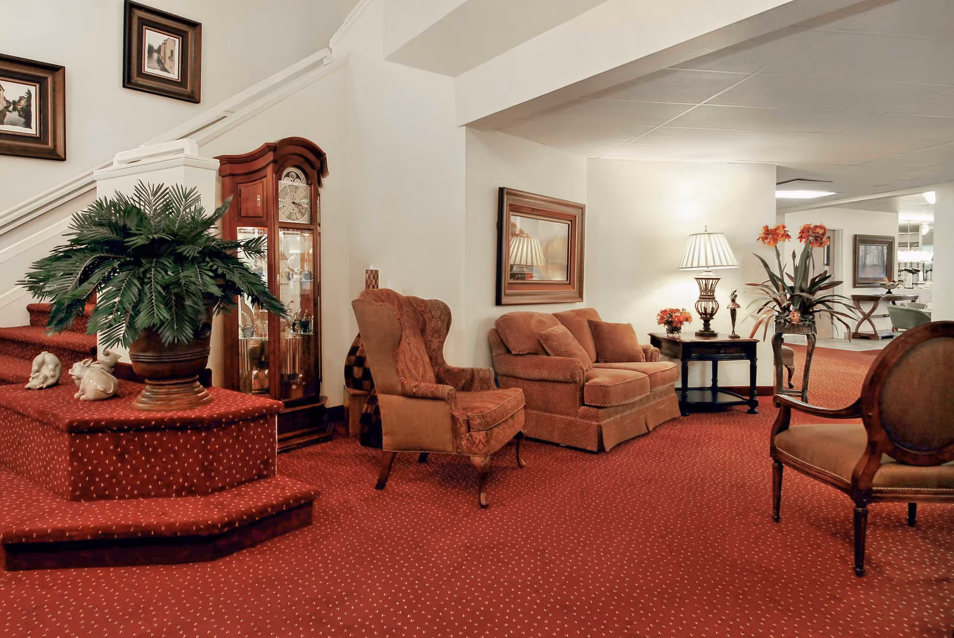 A cozy living room area with red carpet and matching upholstered furniture including an armchair and a sofa. There is a wooden grandfather clock near a staircase with red carpeted steps. A large potted plant sits on a step, and a side table with a lamp and flower arrangement is next to the sofa. The walls are decorated with framed artwork.