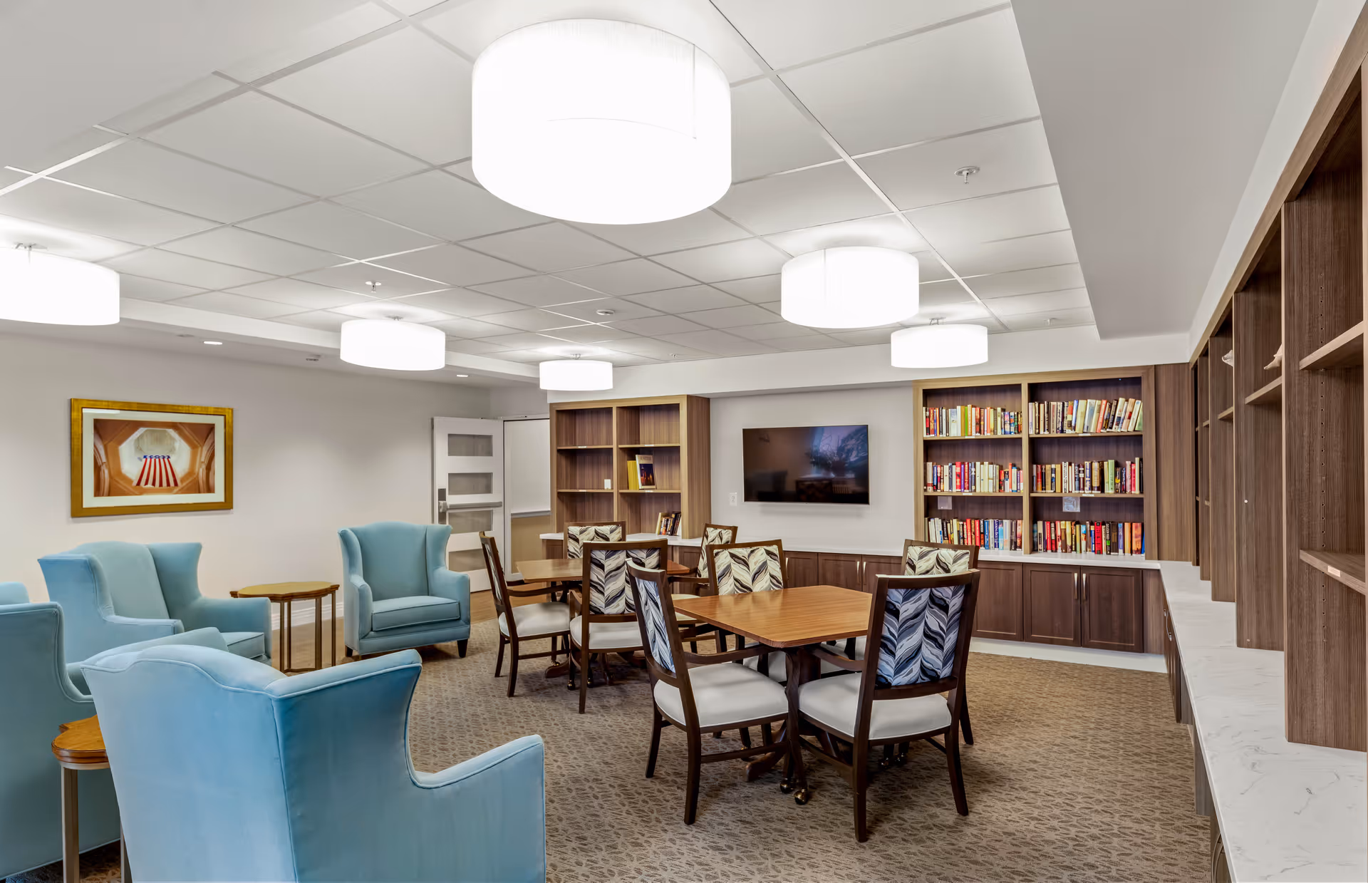 A well-lit common room with several light blue armchairs arranged around small wooden tables, two wooden tables with patterned chairs, bookshelves filled with books, a wall-mounted flat-screen TV, and a framed picture on the wall.