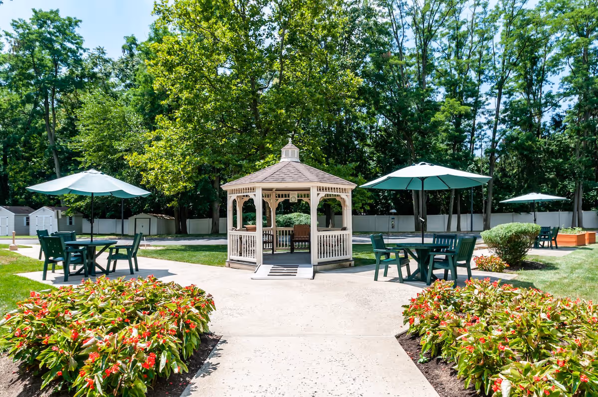 Outdoor garden area with a white gazebo in the center, surrounded by green trees and bushes. There are several green tables with matching chairs and large green umbrellas providing shade. Flower beds with red flowers line the paved walkway leading to the gazebo.
