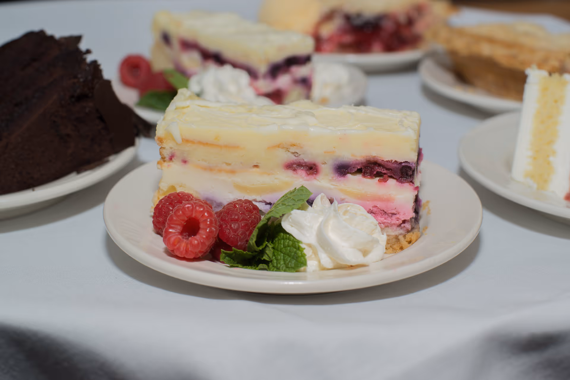 Close-up of a slice of layered berry cheesecake garnished with fresh raspberries, mint leaves, and whipped cream on a white plate, with other dessert slices blurred in the background on a white tablecloth.