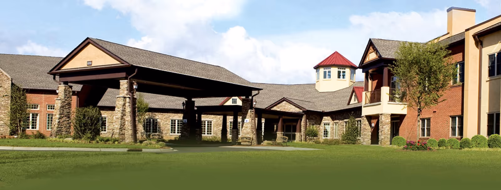 Front exterior of a brick-and-stone senior living building featuring a covered porte-cochere, turret, and manicured lawn.