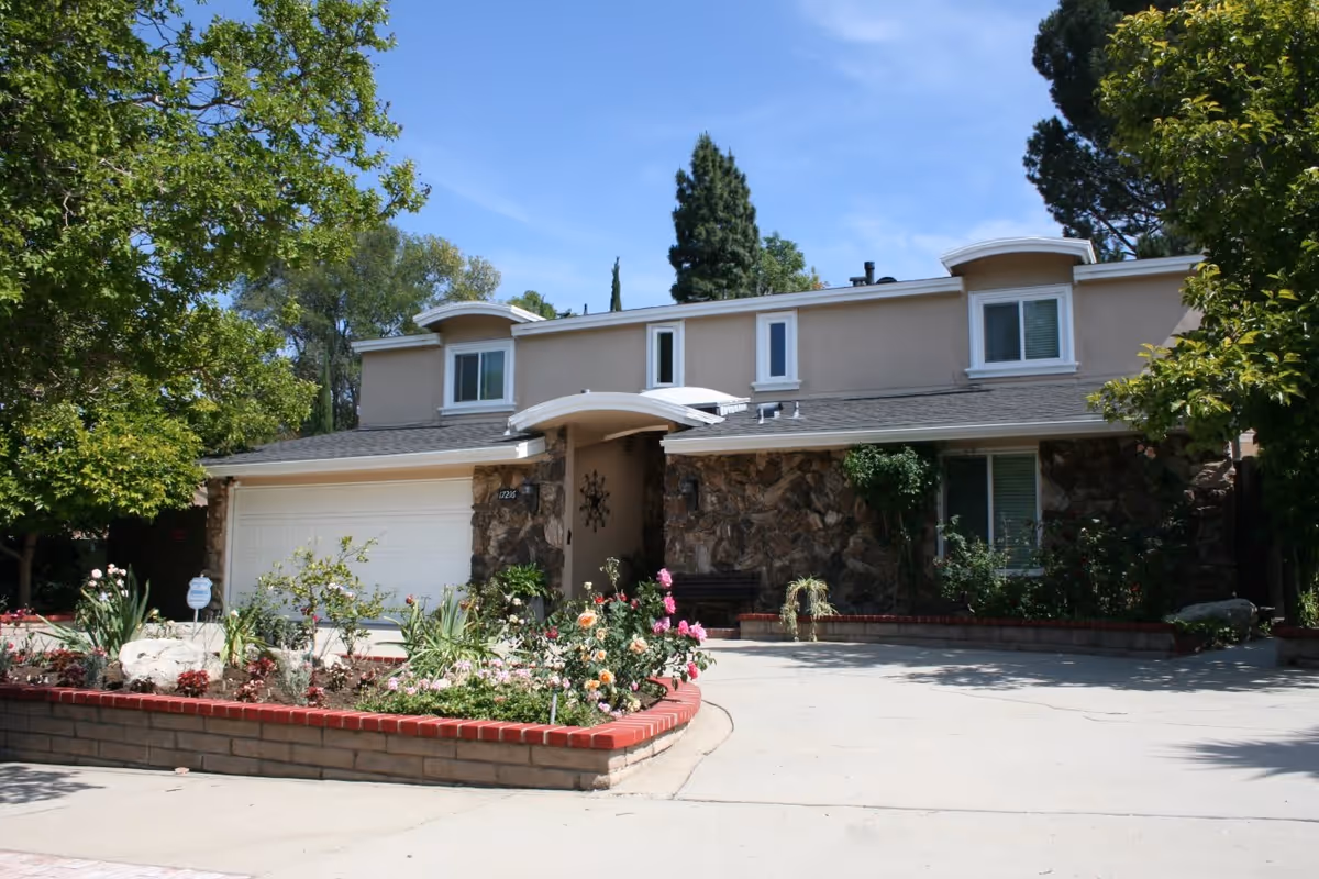 Front exterior view of a two-story residential building with a stone facade on the lower level and beige siding on the upper level. The house has a white garage door, several windows, and a small covered entrance. There is a garden bed with various flowers and plants bordered by red bricks in front of the house, and trees surround the property under a clear blue sky.