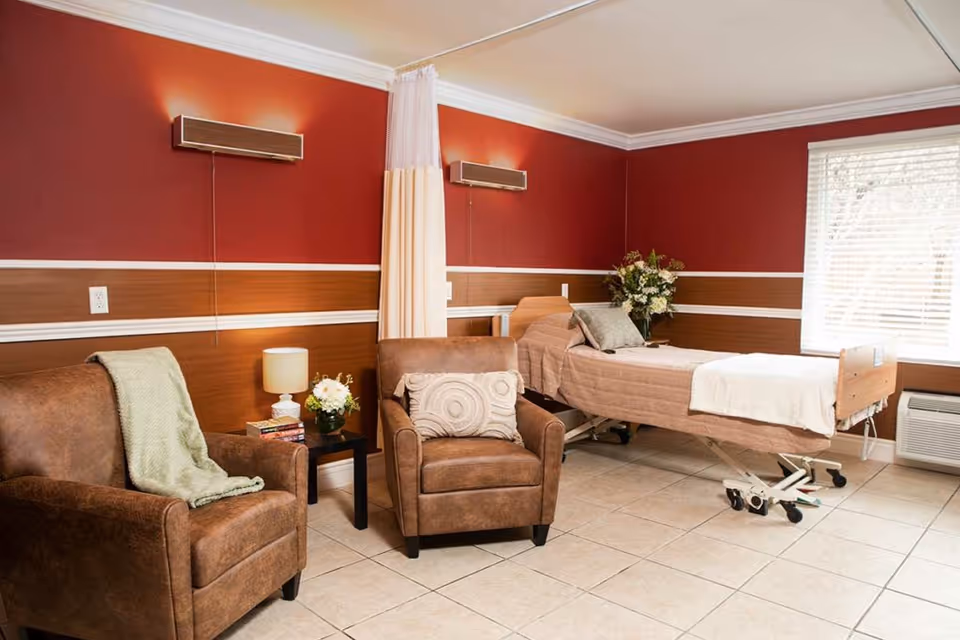 A cozy senior care room with an adjustable bed, two brown armchairs, a side table with lamp and flowers, and red and wood-accented walls.