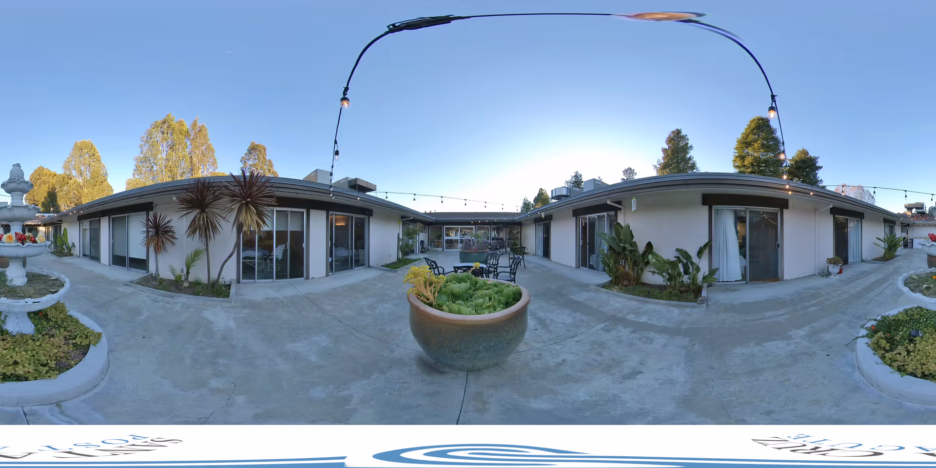 Outdoor central courtyard of a single-story assisted living facility with sliding glass doors, potted plants, a fountain, and string lights.