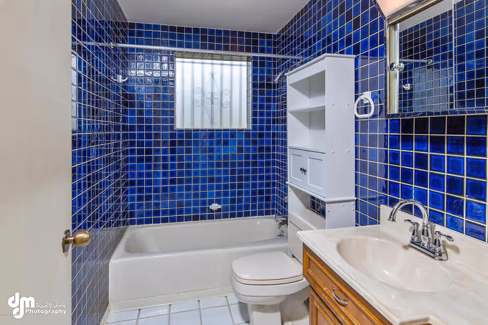 Bathroom with dark blue square tiles on the walls, a white bathtub, a white toilet, a wooden vanity with a beige countertop and sink, a wall-mounted white cabinet with shelves and doors, a mirror above the sink, and a frosted window with bars.