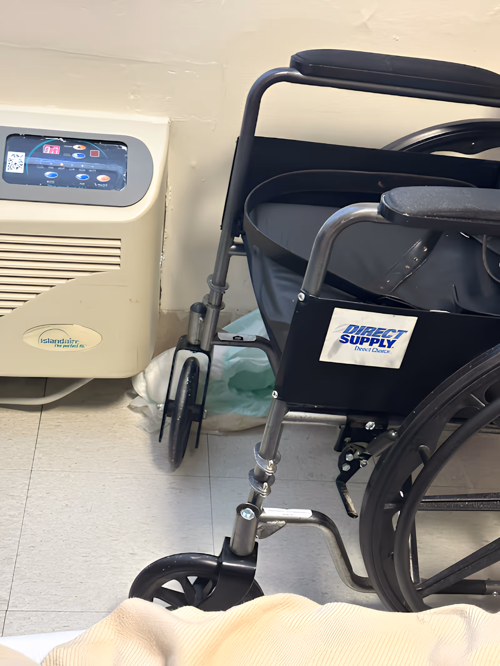 Close-up view of a black wheelchair with a 'Direct Supply' label on the side, positioned next to a beige medical device on a tiled floor, with part of a beige blanket visible in the foreground.