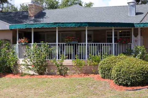 Front porch of a single-story senior living facility with railing, awning, and greenery.