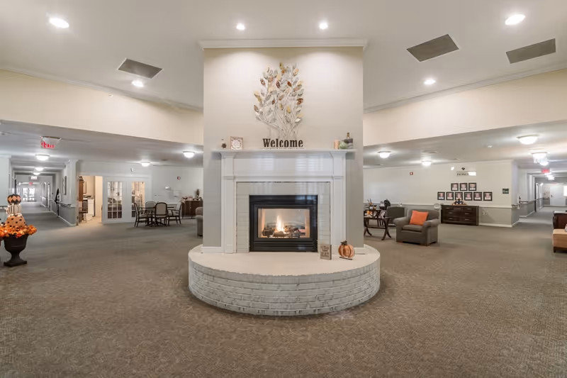 A spacious senior living facility common area with a central white brick fireplace that has a lit fire. Above the fireplace is a decorative metal wall art of leaves and a 'Welcome' sign. The area is carpeted and has seating arrangements with armchairs and tables on either side. The space is well-lit with ceiling lights and has hallways extending to the left and right. Seasonal fall decorations, including pumpkins, are visible near the fireplace.