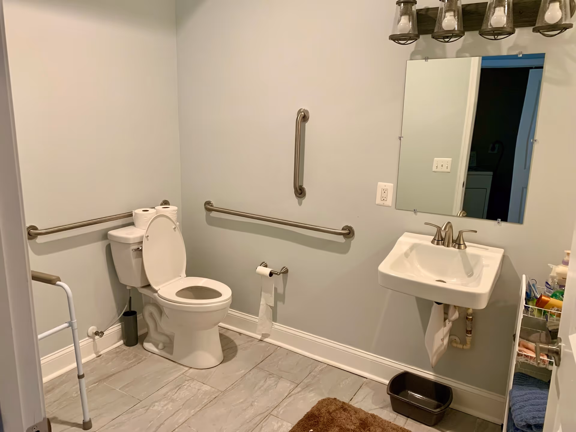 Accessible bathroom with a toilet (lid up), wall-mounted sink and mirror, multiple grab bars, and a walker.