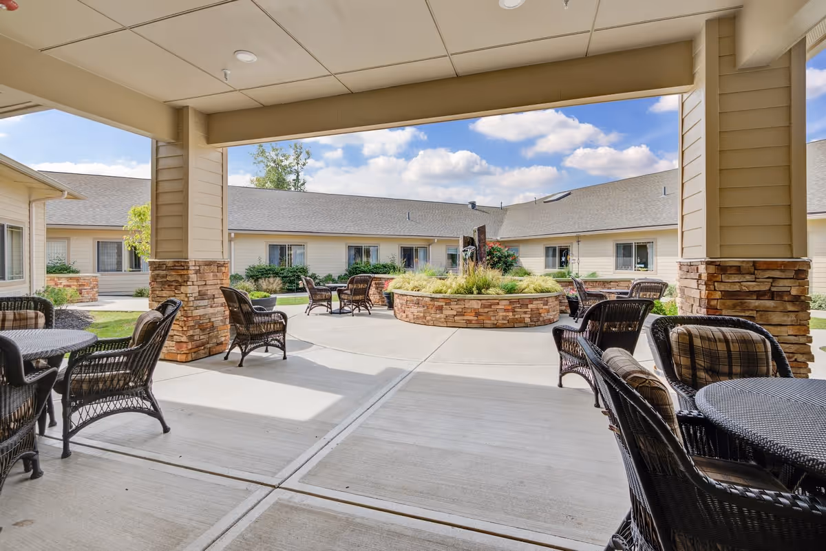 Covered outdoor patio area with wicker chairs and tables, overlooking a courtyard with a circular stone planter filled with greenery and surrounded by a single-story building under a partly cloudy sky.
