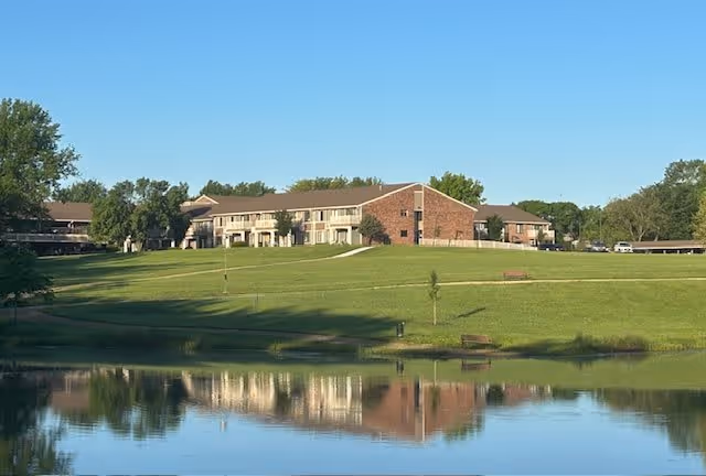 A large senior living facility building with a brick and beige exterior situated on a grassy hill. In the foreground, there is a calm pond reflecting the building and surrounding greenery. The sky is clear and blue.