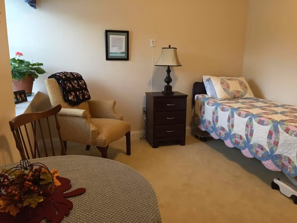 A cozy bedroom in a senior living facility featuring a single bed with a colorful quilt, a wooden nightstand with a lamp, a comfortable upholstered armchair with a black and red blanket draped over it, a wooden chair, a round table with a decorative centerpiece, and a potted plant on the windowsill.