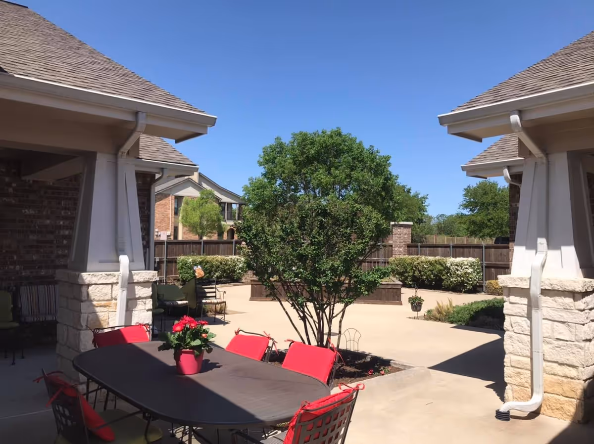 Outdoor patio area with a table and red cushioned chairs under a covered structure. There is a potted plant with red flowers on the table. The patio is surrounded by greenery including a tree and bushes, with a clear blue sky overhead.