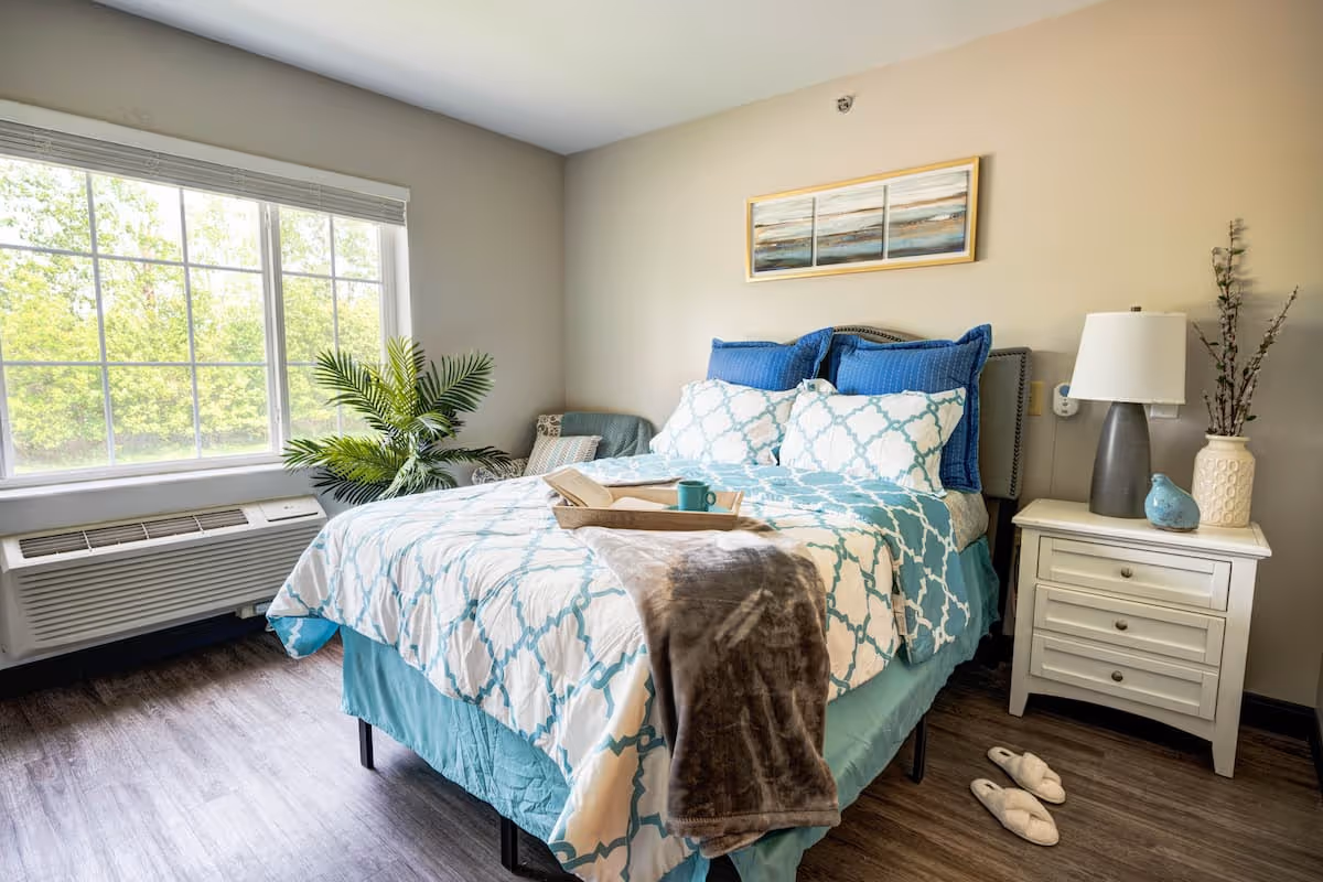 A bright bedroom with a large window showing green trees outside. The bed is neatly made with blue and white patterned bedding, blue pillows, and a gray throw blanket. A wooden tray with a cup and a book rests on the bed. Next to the bed is a white nightstand with a lamp, a vase with branches, and a small blue bird decoration. A pair of white slippers is on the wooden floor beside the nightstand. There is also a green plant near the window.