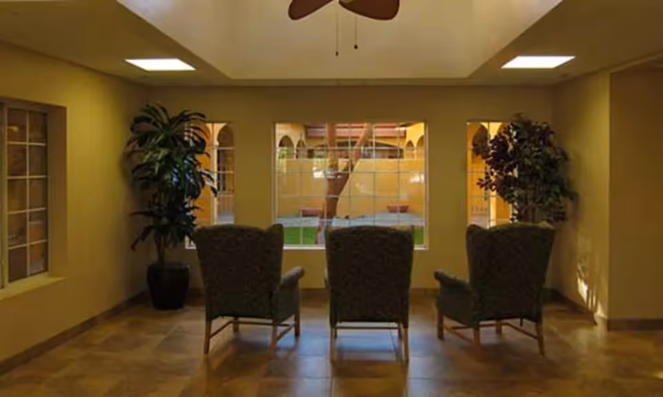 A quiet sitting area with three upholstered armchairs facing a large window that looks out onto a courtyard with a tree and archways. There are two potted plants on either side of the chairs, a ceiling fan above, and tiled flooring.