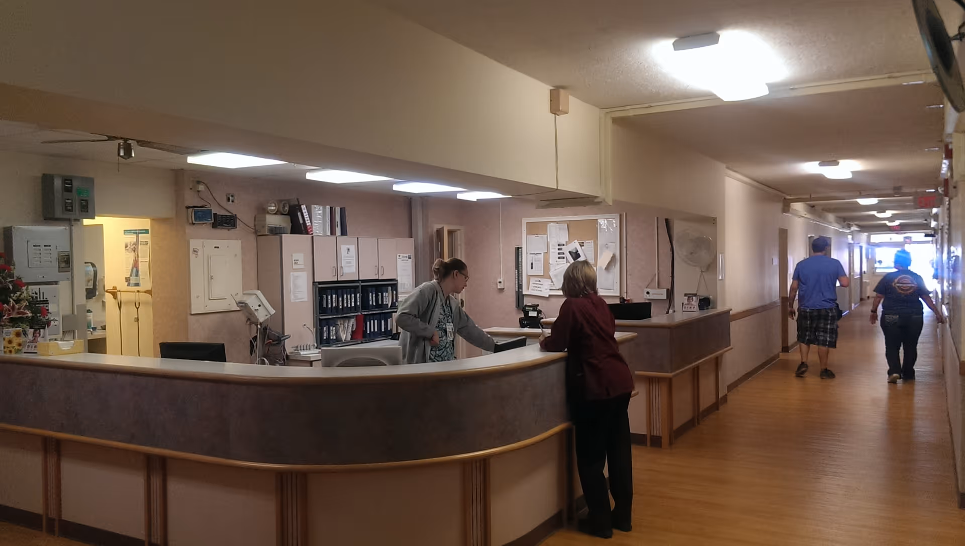 Interior view of a hospital or care facility reception area with a curved desk where a staff member is assisting a visitor. The hallway extends to the right with two people walking away from the camera. The area is well-lit with ceiling lights and has wooden flooring.