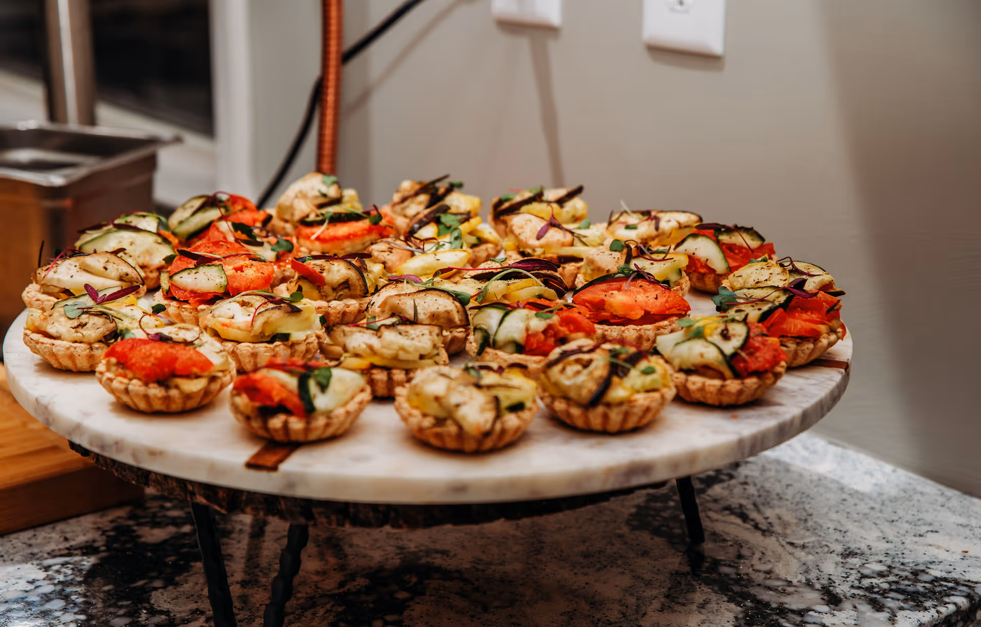 A marble serving platter filled with small tartlets topped with various grilled vegetables including zucchini, tomatoes, and eggplant, placed on a granite countertop in a kitchen setting.
