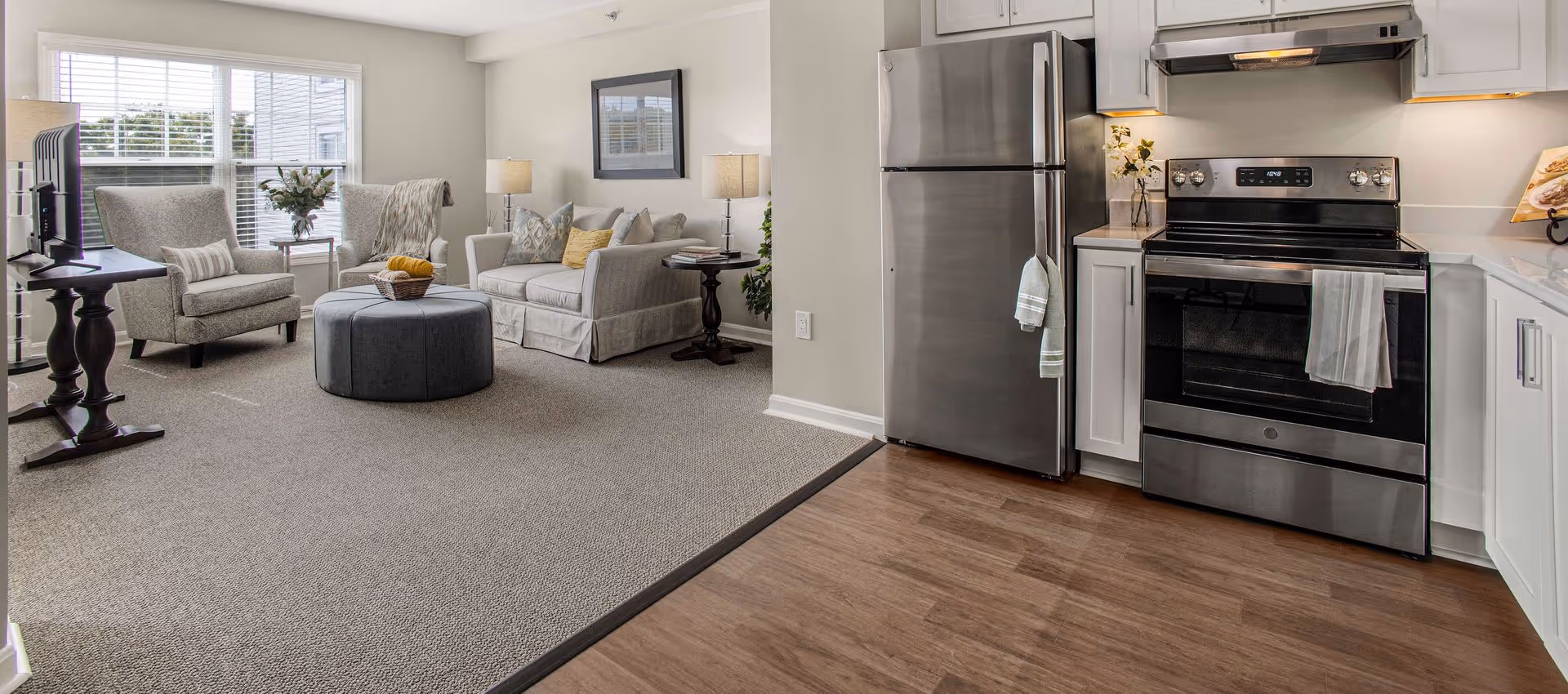A bright and modern living room and kitchen area in a senior living facility. The living room features two armchairs, a sofa with cushions, a round ottoman, side tables with lamps, and a TV on a stand. The kitchen has stainless steel appliances including a refrigerator and an oven, white cabinetry, and a wooden floor transitioning to carpet in the living area.