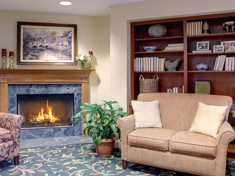 A cozy living room area featuring a lit fireplace with a wooden mantel, a framed painting above it, a potted plant on the floor, a beige loveseat with two light-colored pillows, a floral-patterned armchair, and a wooden bookshelf filled with books, decorative items, and framed photos.
