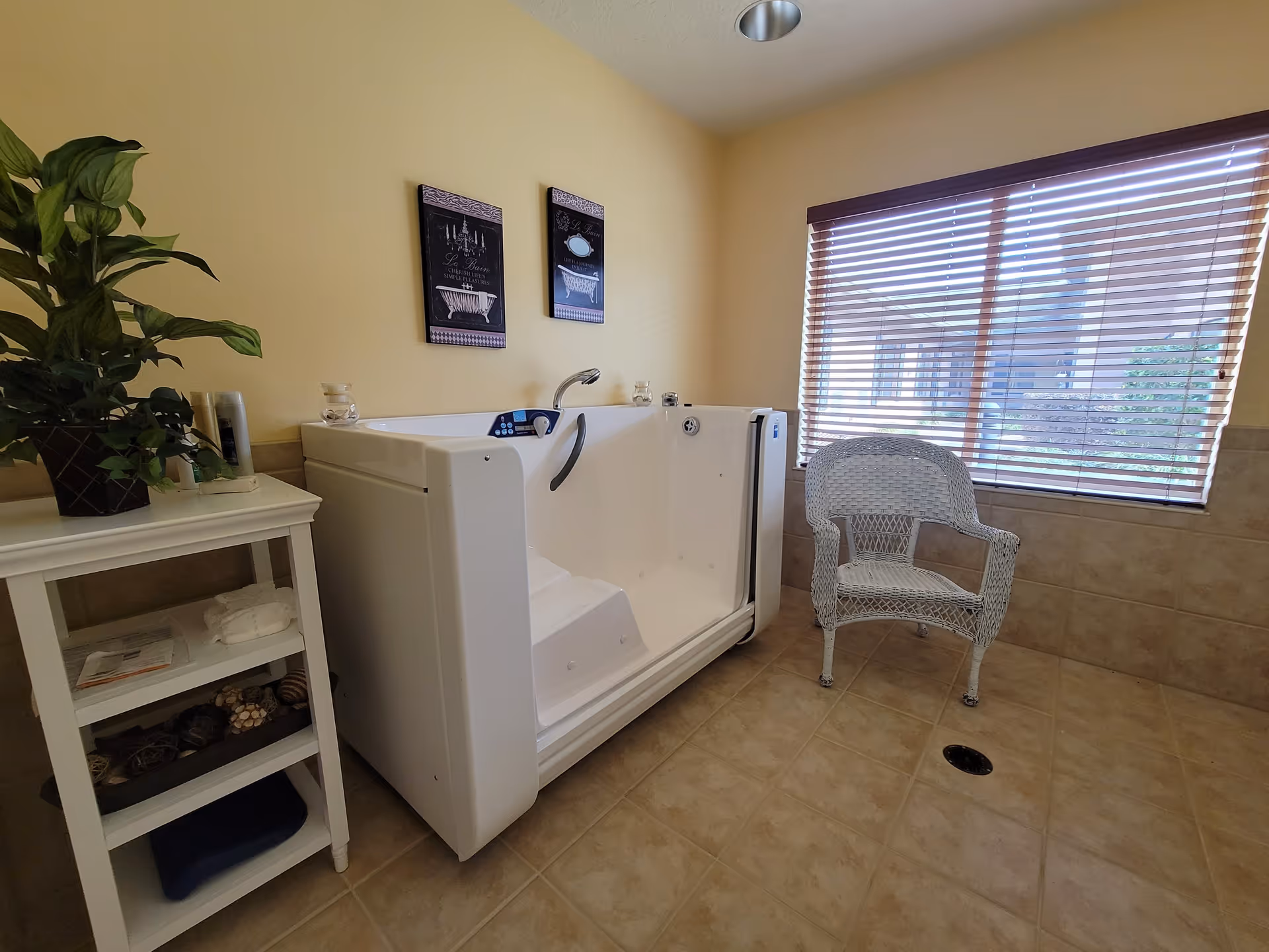 A bright bathroom with a walk-in bathtub, a white wicker chair near a window with wooden blinds, a small white shelf with towels and decorative items, and two framed pictures on the wall above the bathtub.