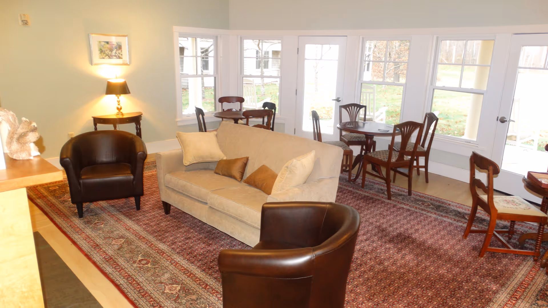 A cozy living room area with a beige sofa adorned with four pillows, two dark brown leather armchairs, and a patterned area rug. Behind the sofa, there are two round wooden tables each surrounded by four wooden chairs with patterned cushions. Large windows and glass doors provide a view of the outdoor greenery and allow natural light to fill the room. A small side table with a lamp and a framed picture on the wall are visible to the left.