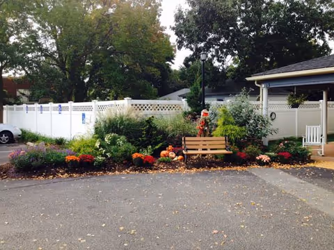 Outdoor garden area with a wooden bench surrounded by various plants and flowers, including orange and purple blooms. A white fence encloses the area, and there is a covered porch to the right. Trees are visible in the background.