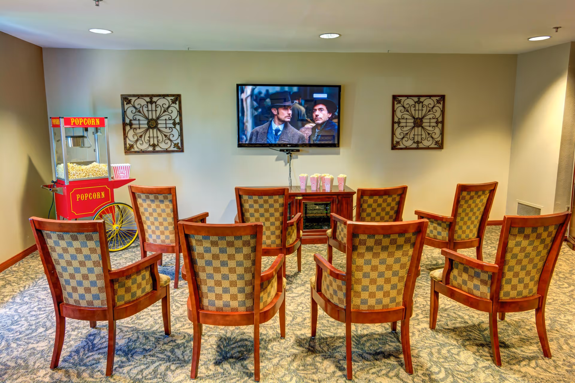 A small theater room with nine wooden chairs arranged in three rows facing a wall-mounted flat screen TV showing a movie scene with two men in hats. To the left of the chairs is a red popcorn machine with popcorn inside and a few popcorn containers on a small wooden cabinet beneath the TV. The room has patterned carpet and two decorative wall hangings on either side of the TV.