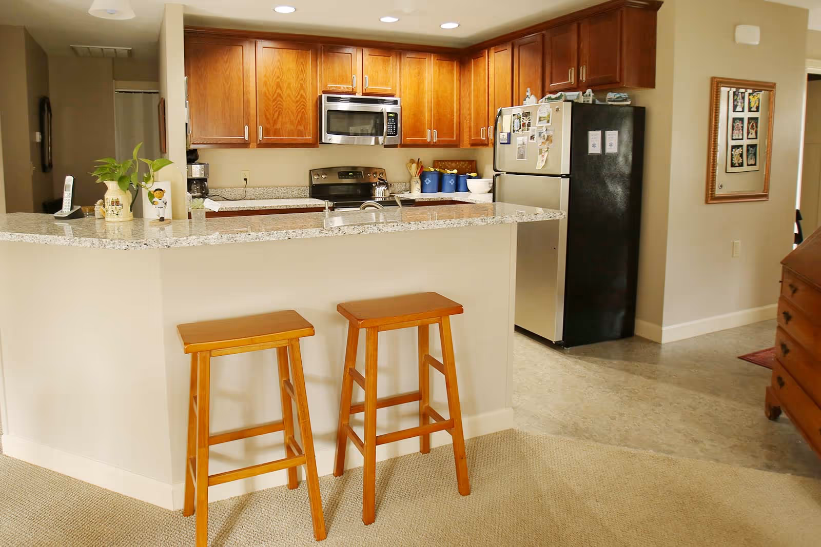 A kitchen area with wooden cabinets, a granite countertop with two wooden stools, a stainless steel microwave and stove, a refrigerator with magnets and notes, and various kitchen items on the counter.