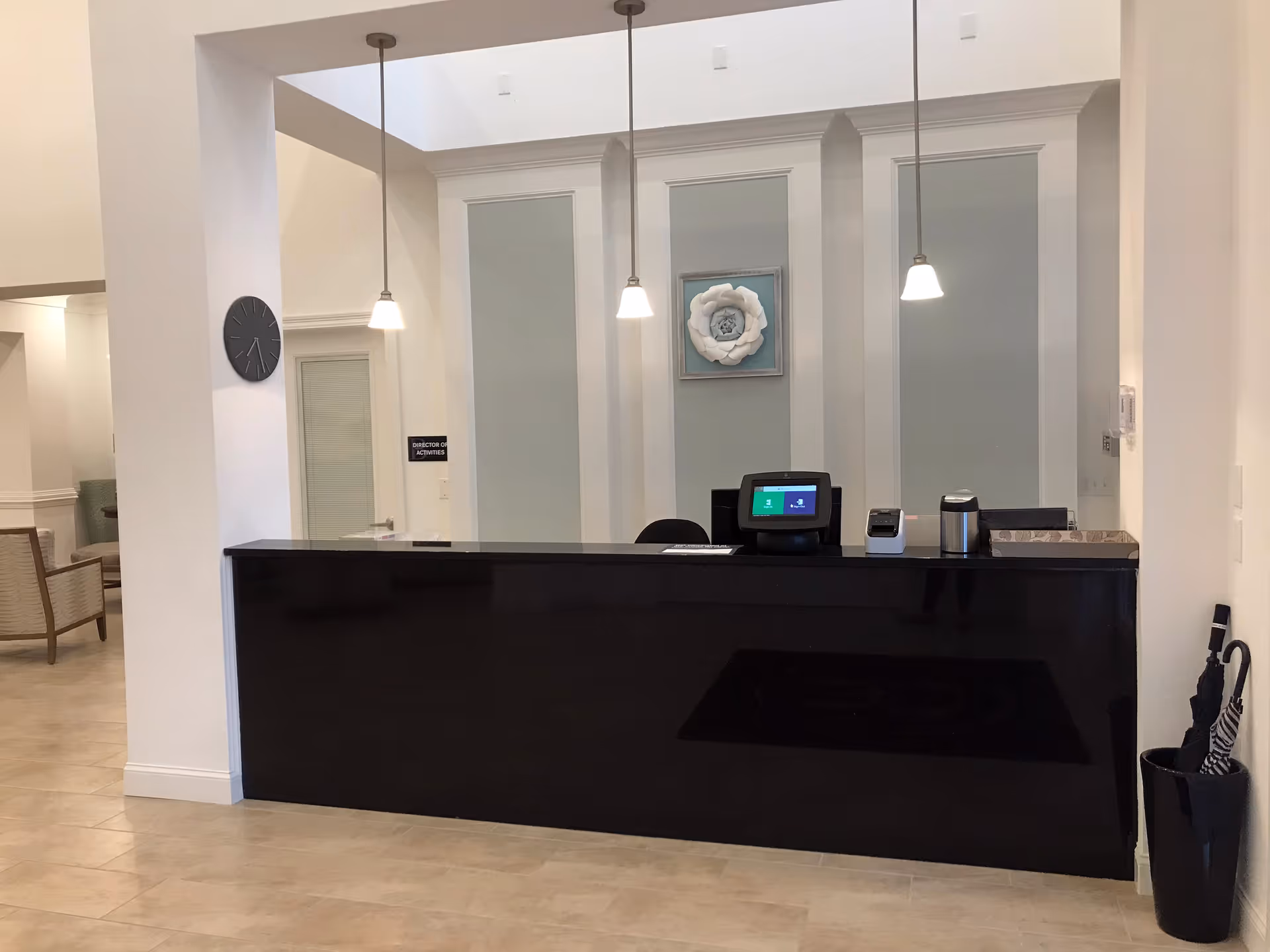 Reception desk area with a black counter, three hanging pendant lights, a clock on the wall, and a decorative flower artwork behind the desk. There is a sign on the wall that reads 'Director of Activities' and an umbrella stand with umbrellas to the right of the desk.