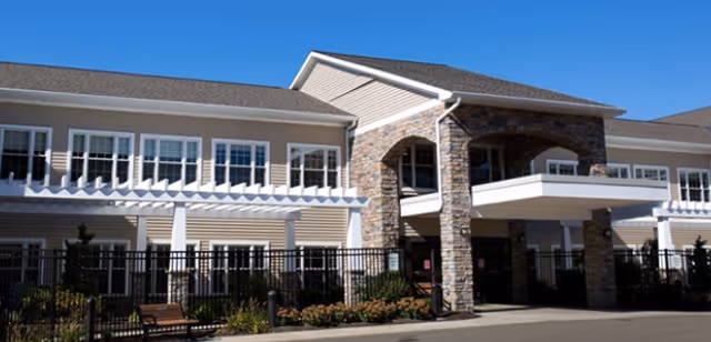 Exterior view of a two-story senior living facility building with beige siding and stone accents around the entrance. The building has multiple windows, a covered entrance, and a black metal fence enclosing a landscaped area with benches and plants. The sky is clear and blue.