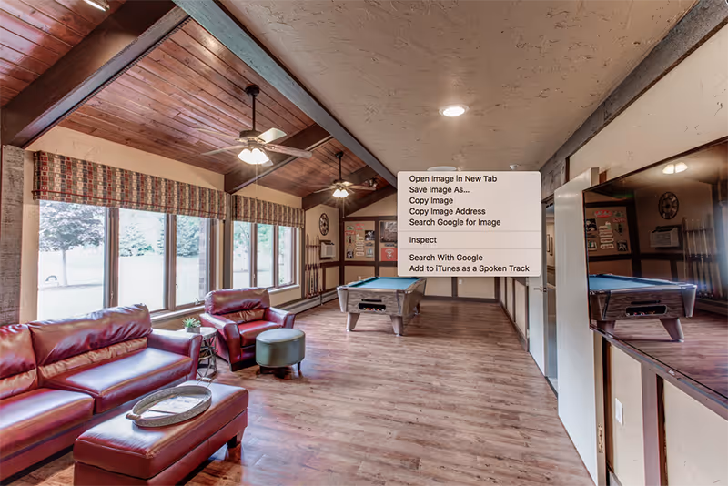 A spacious common area with wooden floors and a high wooden ceiling featuring ceiling fans. The room has large windows with patterned valances letting in natural light. There are red leather sofas and an ottoman arranged near the windows, and a pool table is positioned further back in the room. A large flat-screen TV is mounted on the wall to the right.