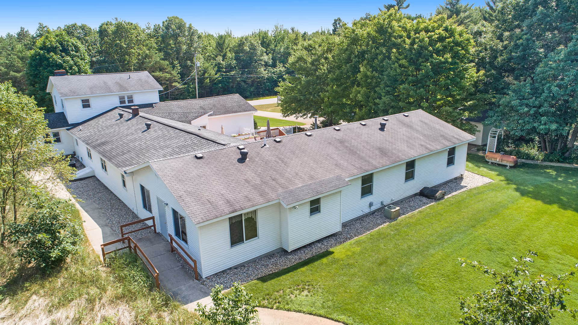 Aerial view of a single-story white building with a gray roof surrounded by green grass and trees, showing the exterior of Pine Ridge Assisted Living facility on a sunny day.