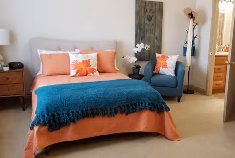 A cozy bedroom featuring a bed with an orange bedspread, matching pillows, and a teal throw blanket. Next to the bed is a wooden nightstand with a lamp and clock. A blue armchair with a floral pillow sits beside a small round table with a white orchid. A coat rack with hats and bags is near the doorway leading to a bathroom.