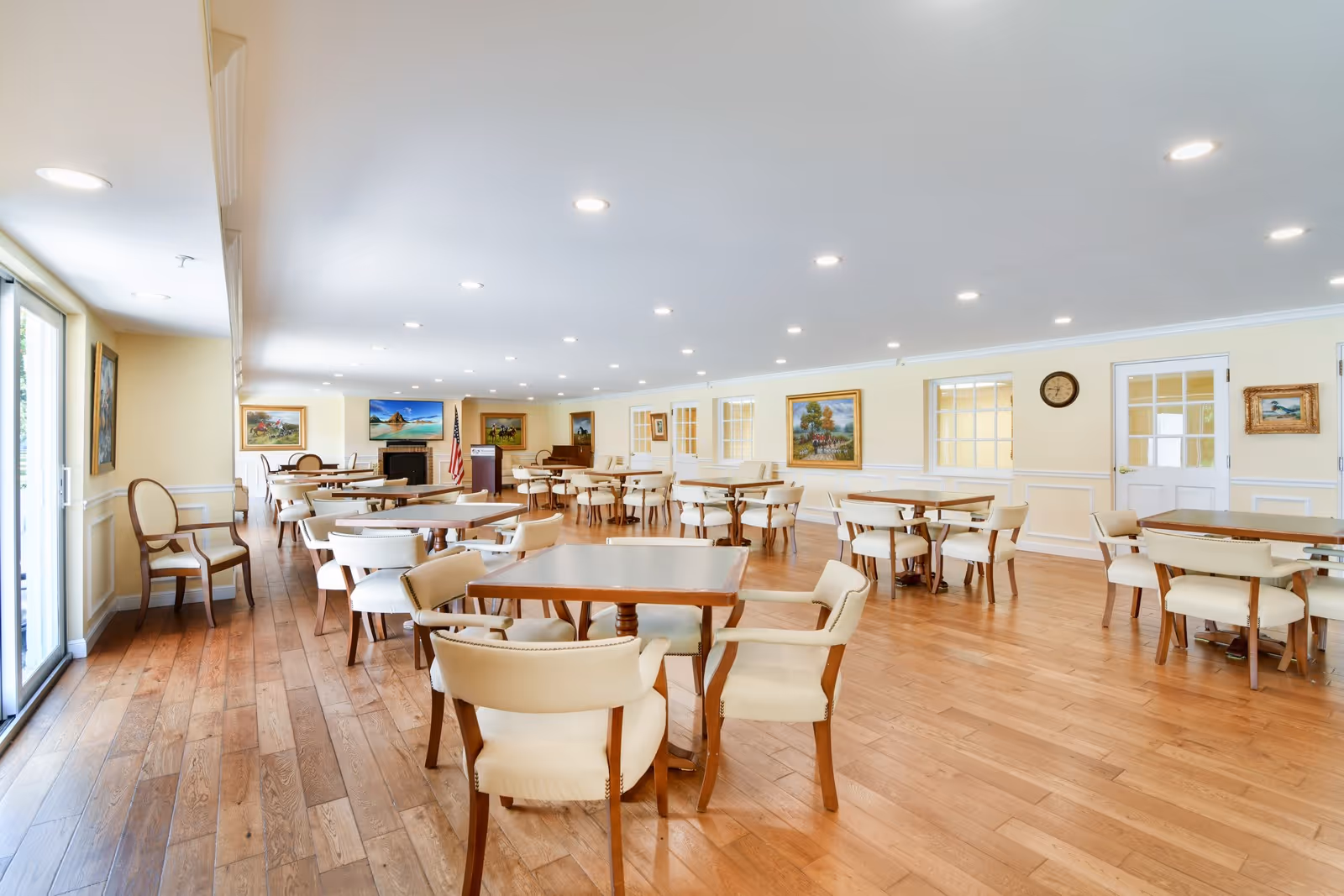 A spacious dining room with multiple wooden tables and cream-colored cushioned chairs arranged neatly on a wooden floor. The room has light yellow walls adorned with framed paintings and a clock. Large windows and glass doors on the left side allow natural light to enter. The ceiling is fitted with numerous recessed lights, and there is a fireplace with an American flag and a podium near the back wall.