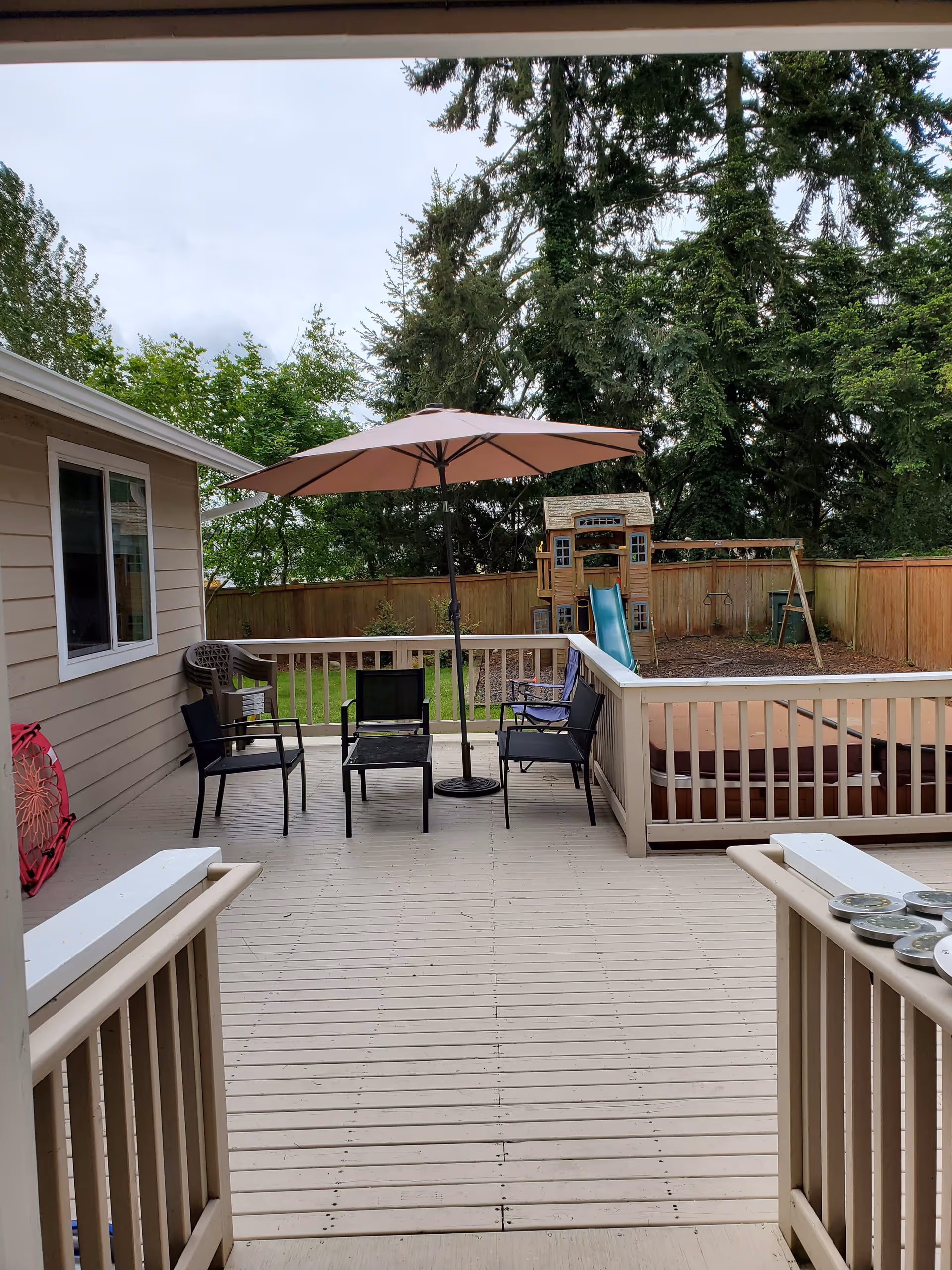 Outdoor wooden deck area with a large umbrella, several chairs, a small table, and a hot tub. In the background, there is a fenced yard with a children's play structure including a slide and swings, surrounded by tall trees.