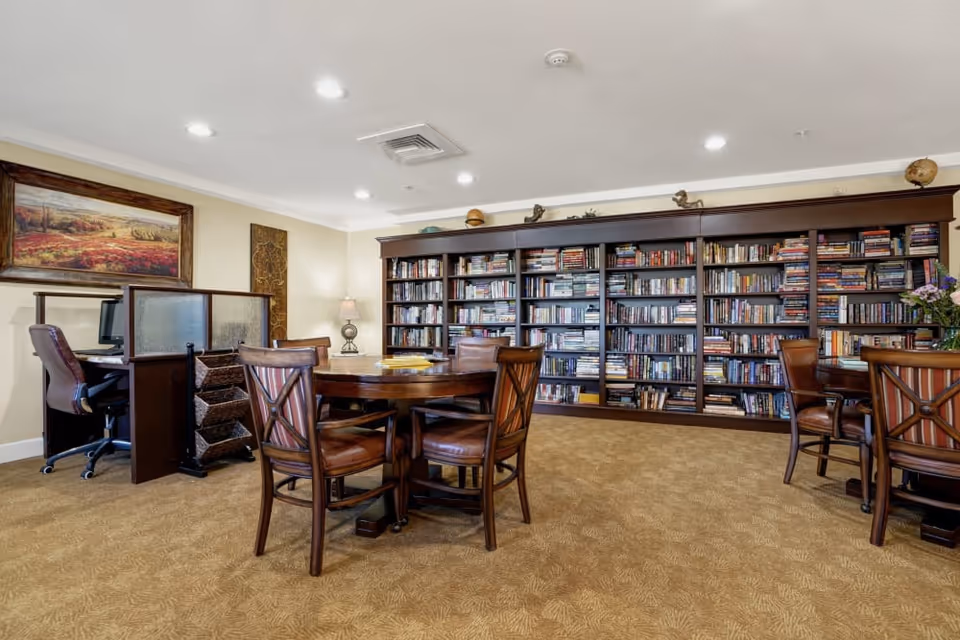 Well-lit library/common room with round wooden tables and chairs, a computer workstation, and a large wall of bookshelves.