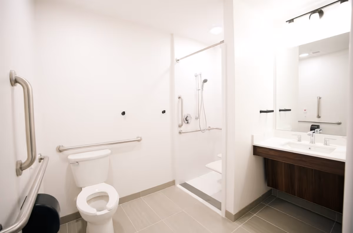 A clean, accessible bathroom with white walls and light gray tiled floor. The room features a toilet with grab bars on the walls, a walk-in shower with a handheld showerhead and multiple grab bars, and a vanity with a large mirror and two faucets.