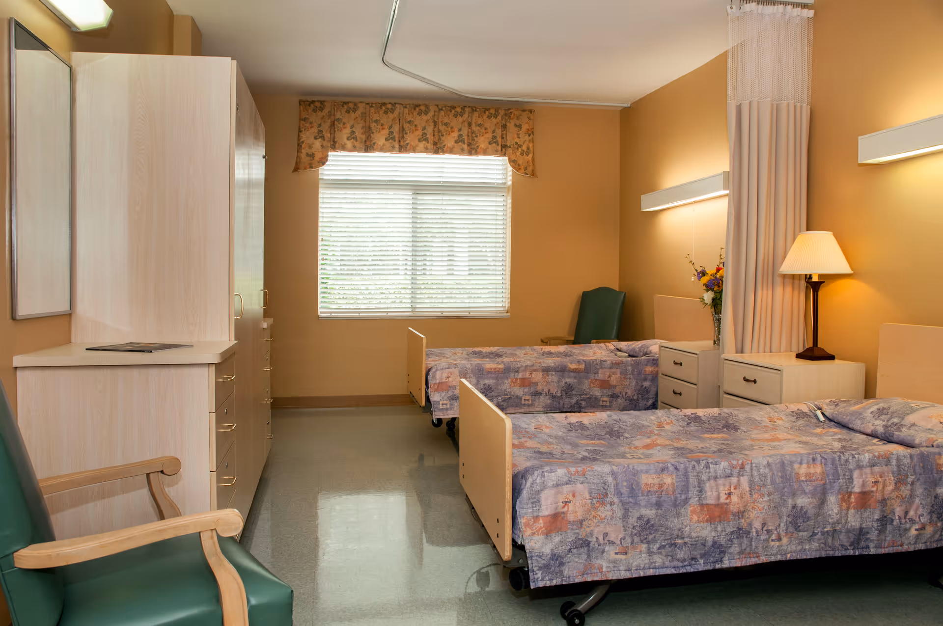 A senior living bedroom with two hospital-style twin beds, bedside tables, a wardrobe, chairs and a window.
