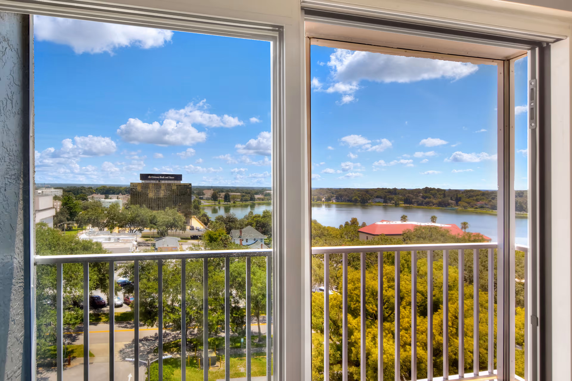 View through a sliding glass door and window showing a balcony railing, green trees, a lake, buildings, and a blue sky with scattered clouds.