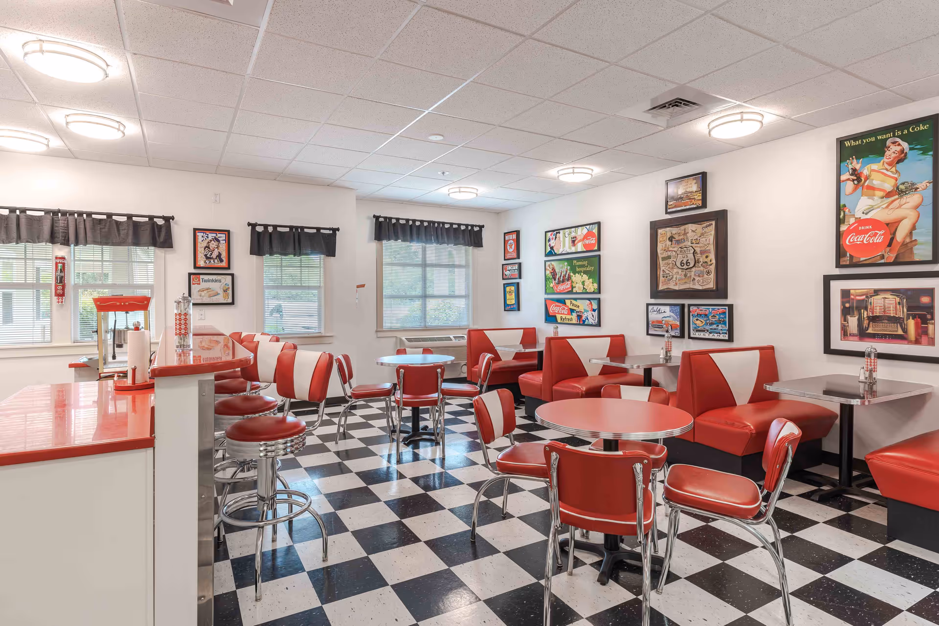 Retro 1950s-style dining room with red-and-white booths, round tables and chrome stools on a black-and-white checkered floor with vintage Coca-Cola decor.