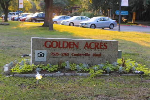 Stone sign for Golden Acres with red lettering displaying the address 2525-2765 Centerville Road, surrounded by small plants and grass with parked cars and trees in the background.