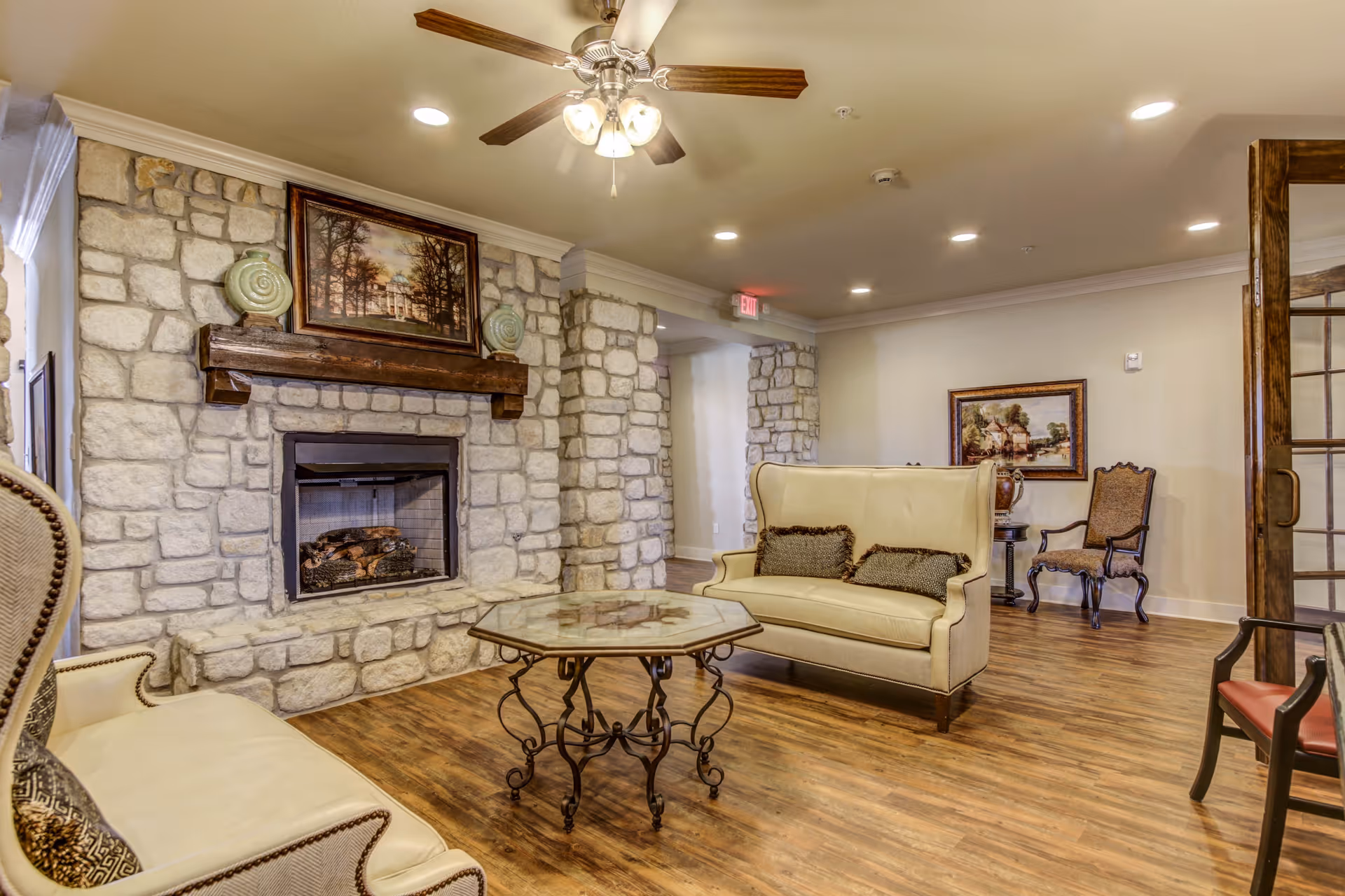 A cozy living room area with a stone fireplace, two cream-colored sofas with decorative pillows, a glass-top coffee table with ornate metal legs, wooden flooring, and framed artwork on the walls. There is a ceiling fan with lights and recessed lighting in the ceiling.
