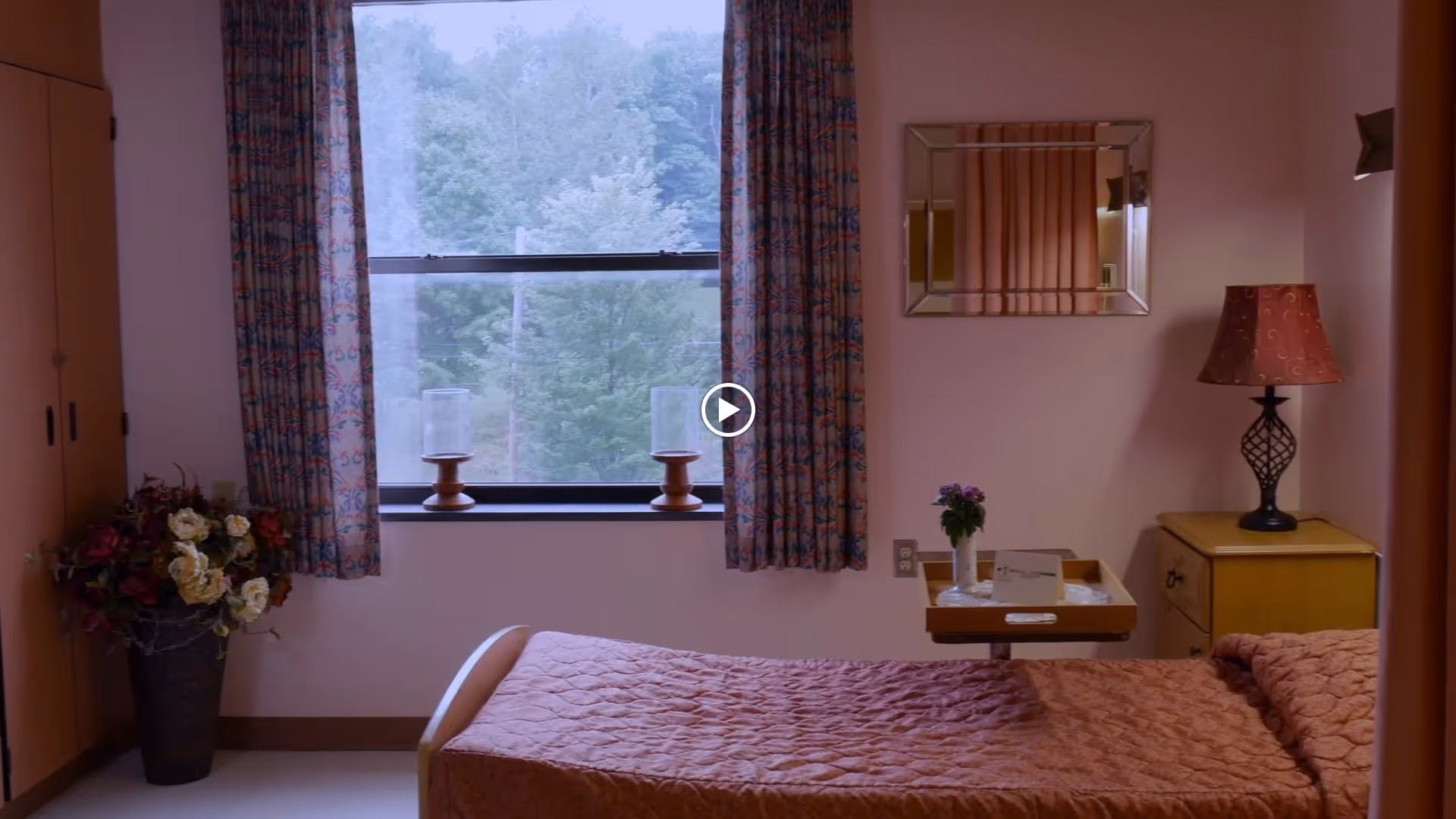 A cozy bedroom in a healthcare facility with a single bed covered in a pink quilted blanket. There is a window with floral curtains showing green trees outside. On the windowsill, there are two candle holders. Next to the bed is a wooden nightstand with a decorative lamp and a small table holding a vase with purple flowers and some papers. A large flower arrangement is placed in a vase on the floor near a closet with wooden doors.