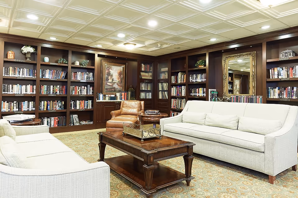 A cozy senior living facility library with built-in dark wood bookshelves filled with books, two light-colored sofas facing each other, a wooden coffee table in the center, a brown leather armchair, a framed painting on the wall, and a large ornate mirror. The room has a patterned carpet and a white coffered ceiling with recessed lighting.