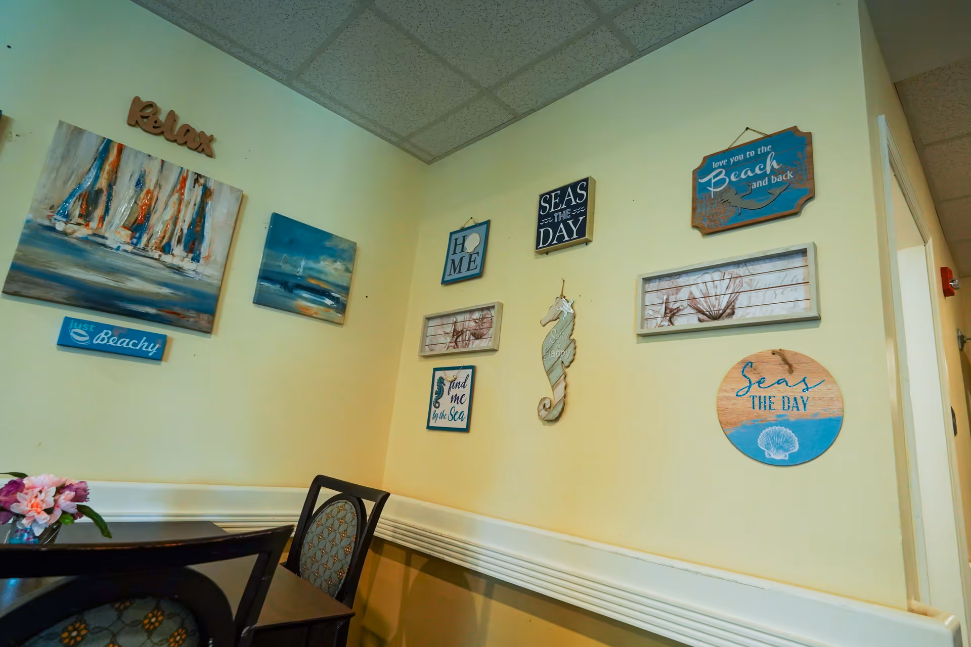Corner of a room with light yellow walls decorated with various beach-themed wall art including paintings, signs, and a seahorse figure. There is a small table with chairs and a vase of flowers in the foreground.